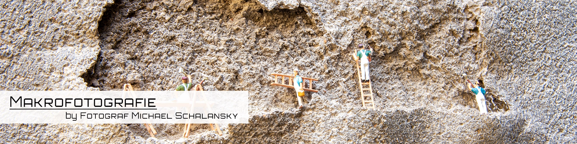 Vier winzige Miniaturfiguren, die auf kleinen Leitern an einer groben, strukturierten Wand scheinbar Putzarbeiten ausführen, dargestellt in Makrofotografie von Michael Schalansky.